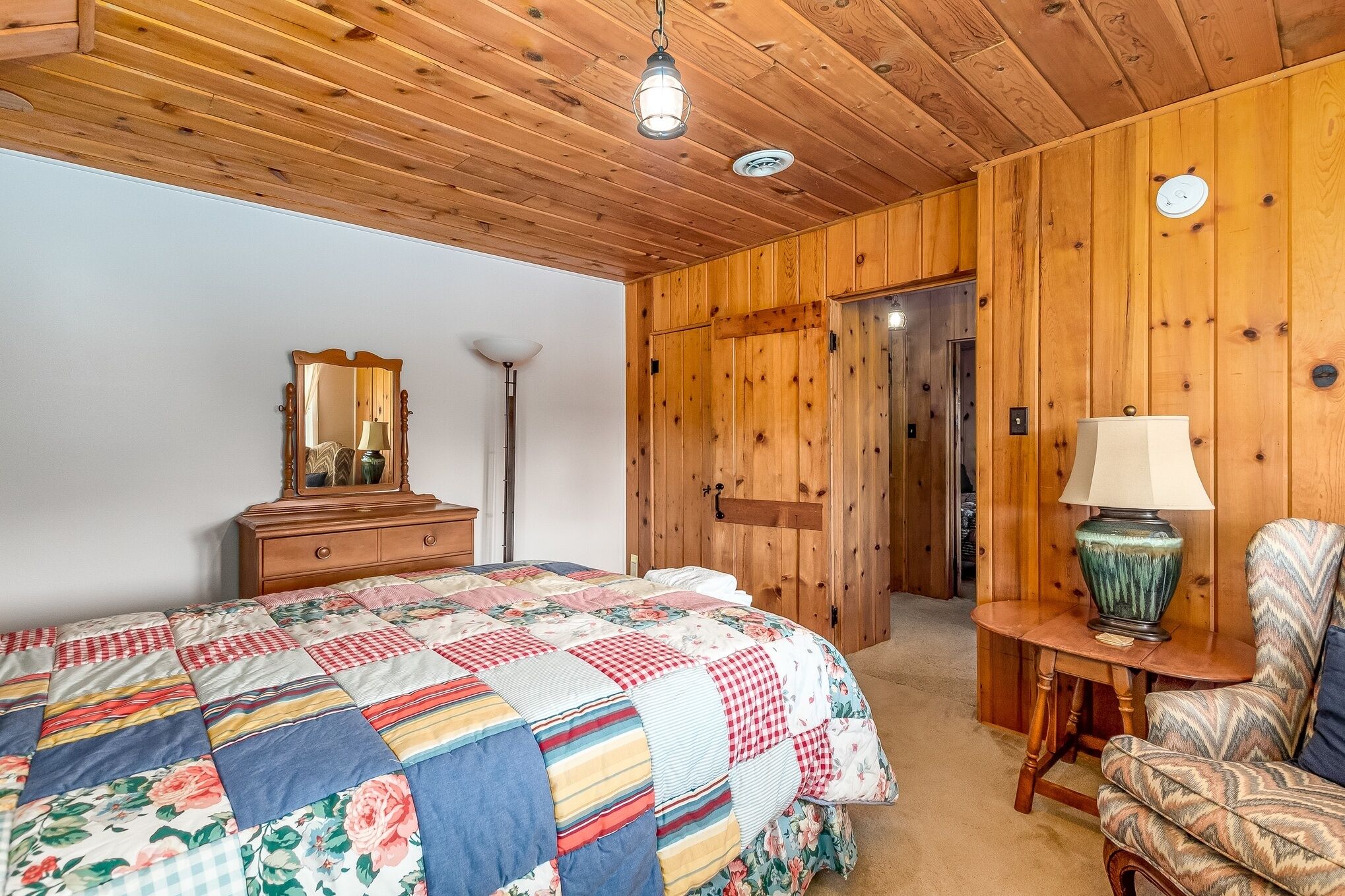 Un dormitorio con techo de madera, cama acolchada, cómoda, espejo, lámpara y una silla.