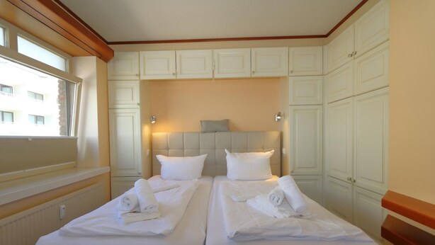 A hotel room with two beds, each with neatly folded towels and pillows, a headboard, and a window with blinds. - App. Hamelmann - with a small sea view (Sylt)