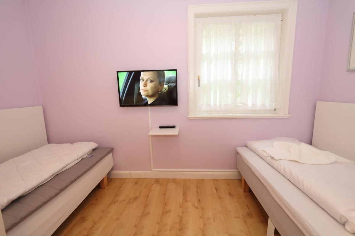 Ein Zimmer mit zwei Betten, einem an der Wand befestigten Fernseher und einem mit Vorhängen versehenen Fenster.