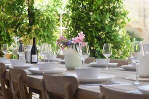 Una mesa dispuesta para una comida, con platos, cuencos y copas de vino blancos, rodeada de follaje.