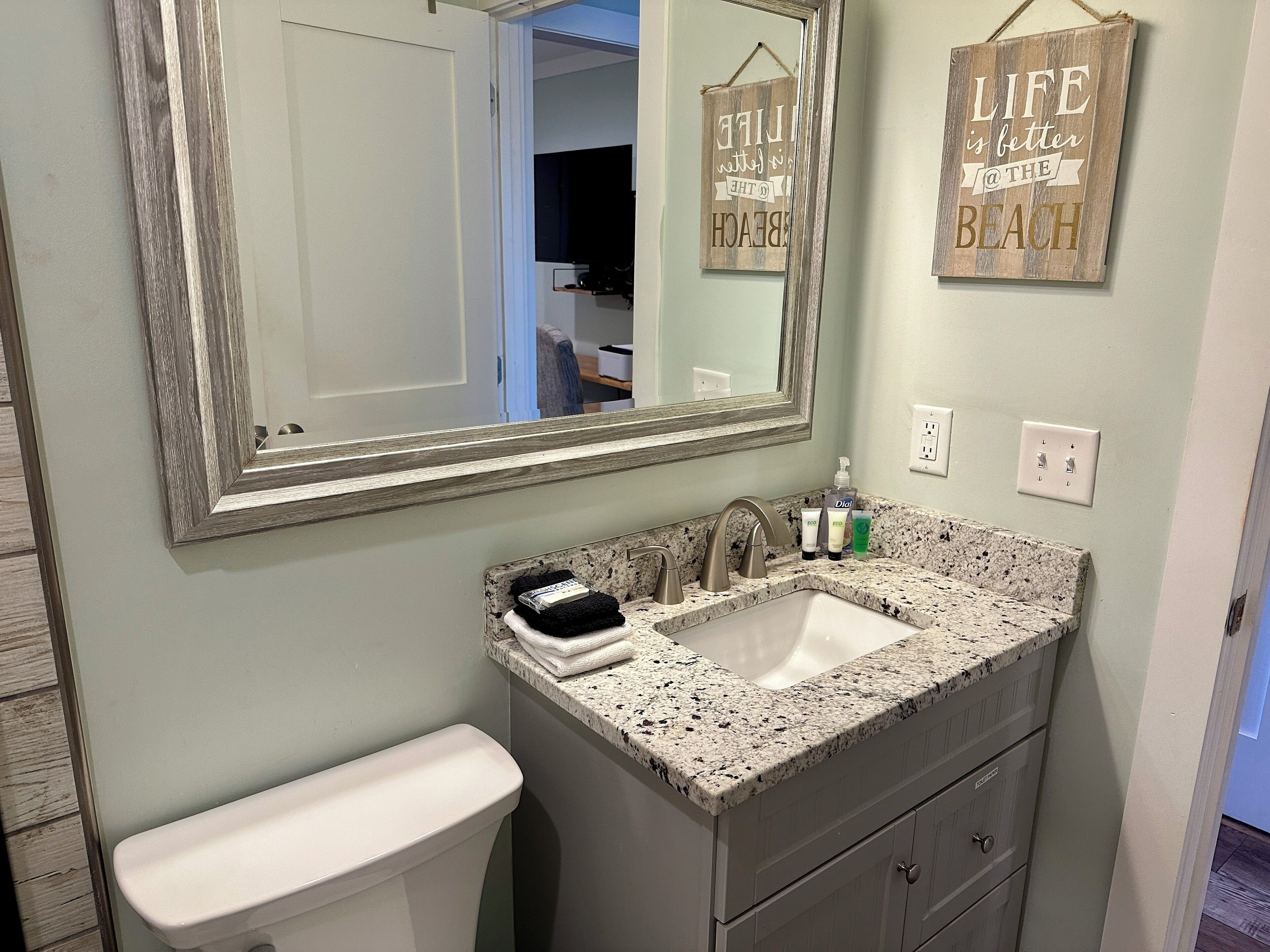 A bathroom with a white toilet, a granite countertop sink, and a large mirror. There are two wall-mounted light switches and a small shelf with toiletries.