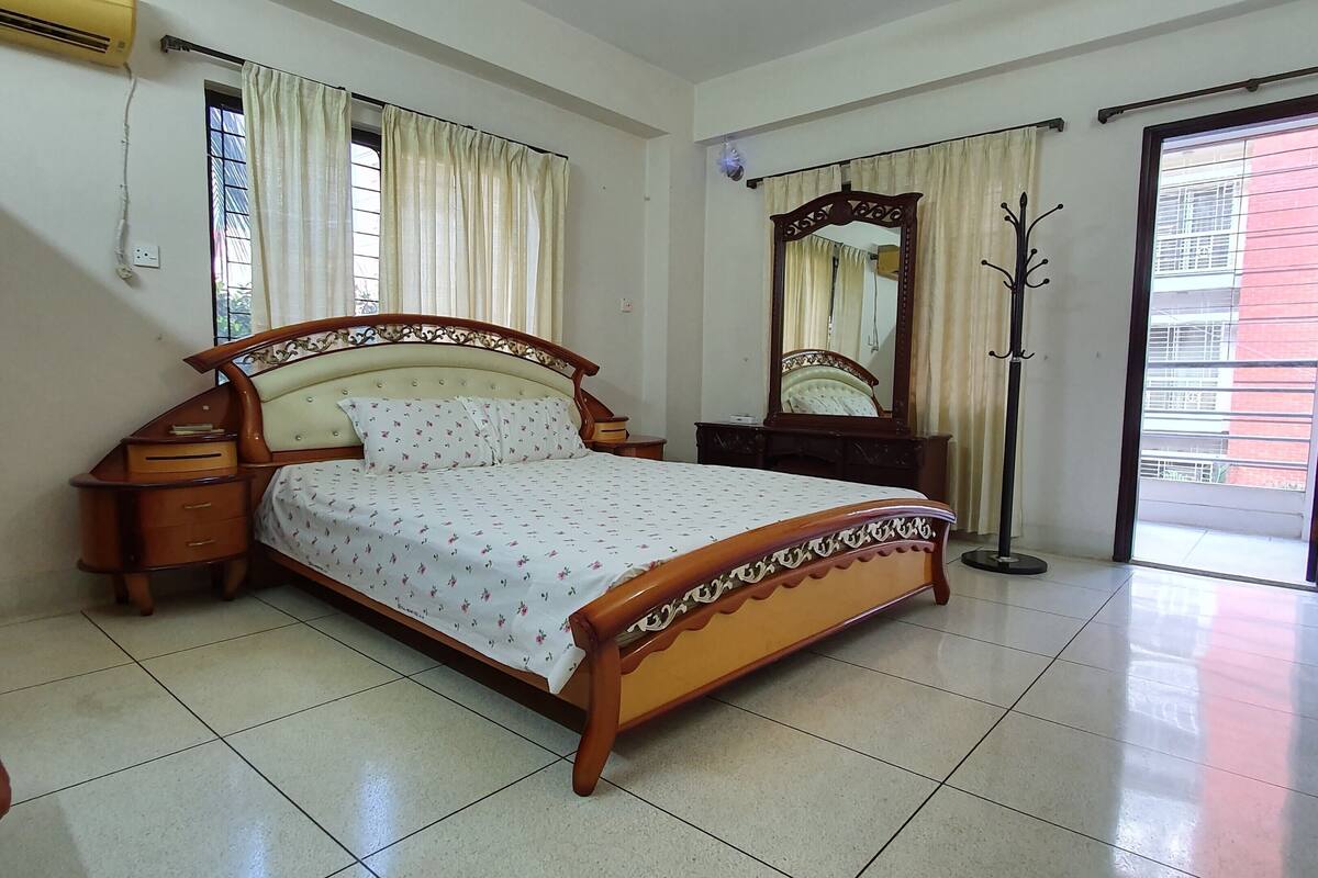 A bedroom with a wooden bed, a nightstand, a dresser, a mirror, and a window with curtains.