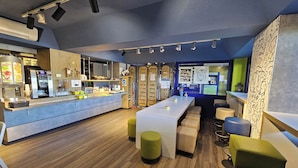 A modern bar area with a long counter, stools, and a seating area with a high ceiling and exposed beams. - B&B Hotel Thionville Yutz A31 (Yutz)