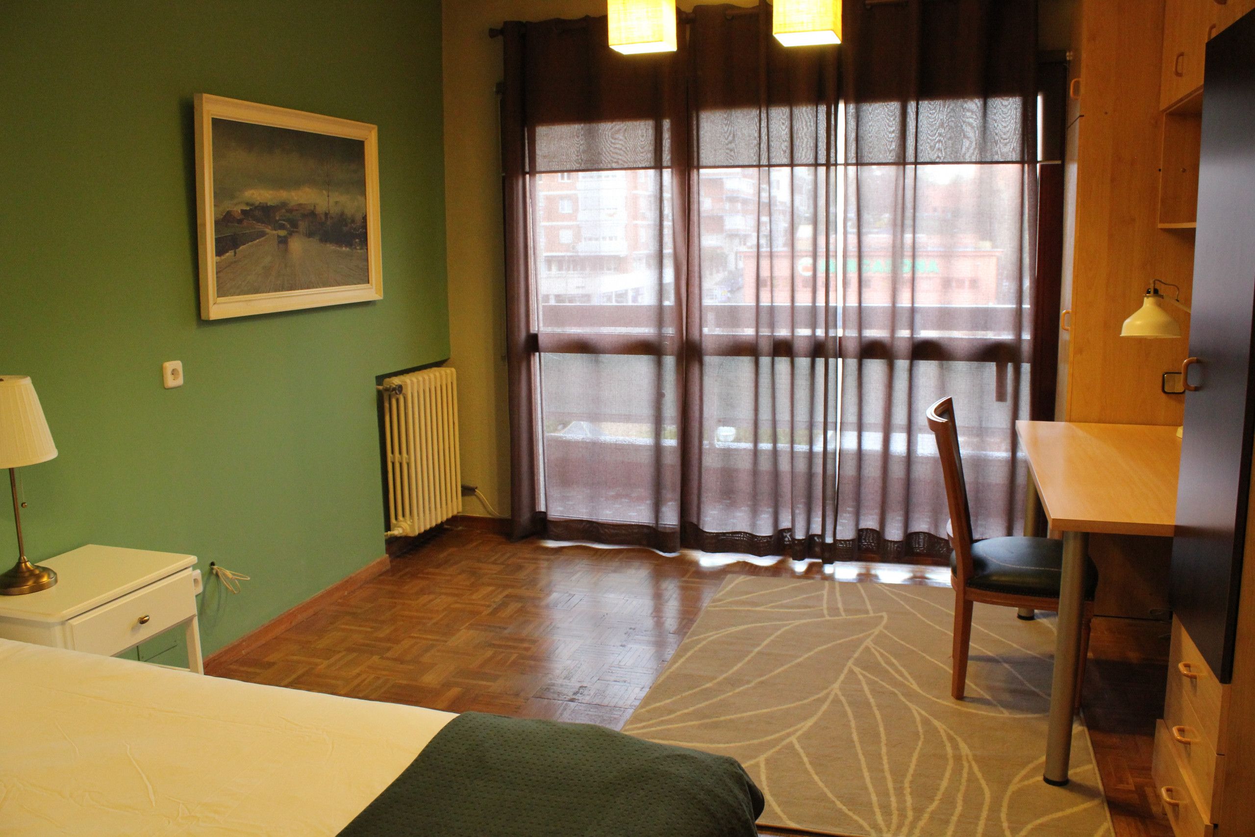 Een hotelkamer met een bed, een stoel, een bureau, een radiator, een raam met gordijnen en een schilderij aan de muur.