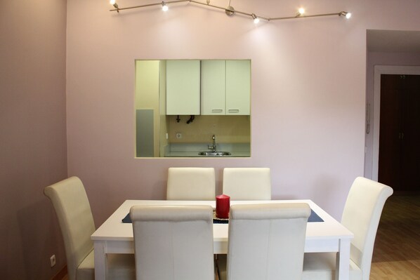Un comedor con una mesa blanca y cuatro sillas, una vela roja y una cocina con armarios verde claro.