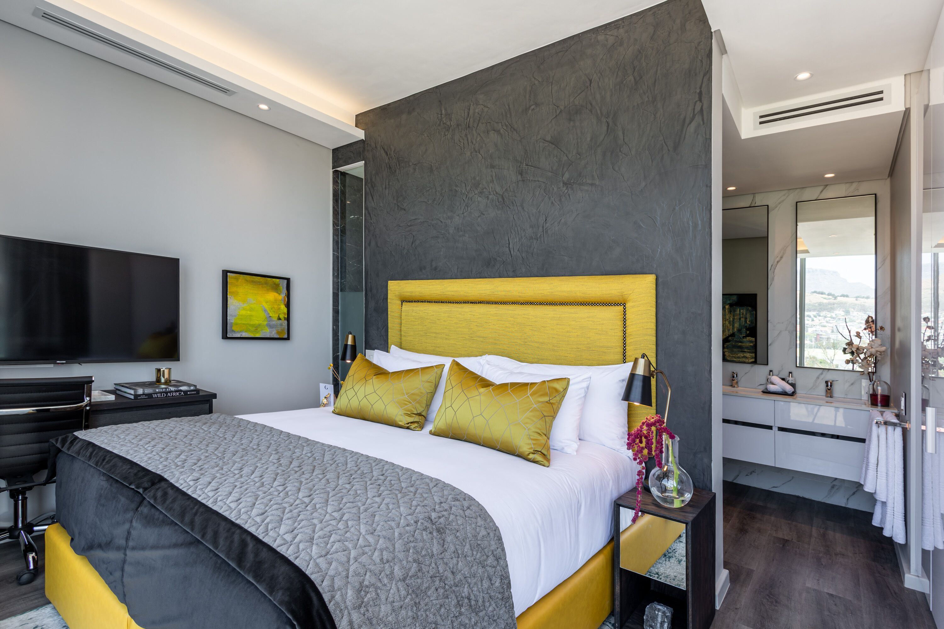 A modern bedroom with a large bed, a flat-screen TV, and a bathroom with a large mirror and white cabinets.