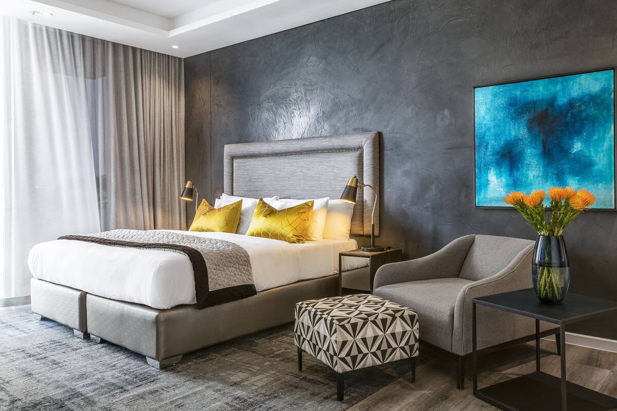 Une chambre d’hôtel moderne avec un grand lit, une table de chevet avec une lampe, une chaise, une petite table ornée d’un vase de fleurs et un grand tableau abstrait accroché au mur.