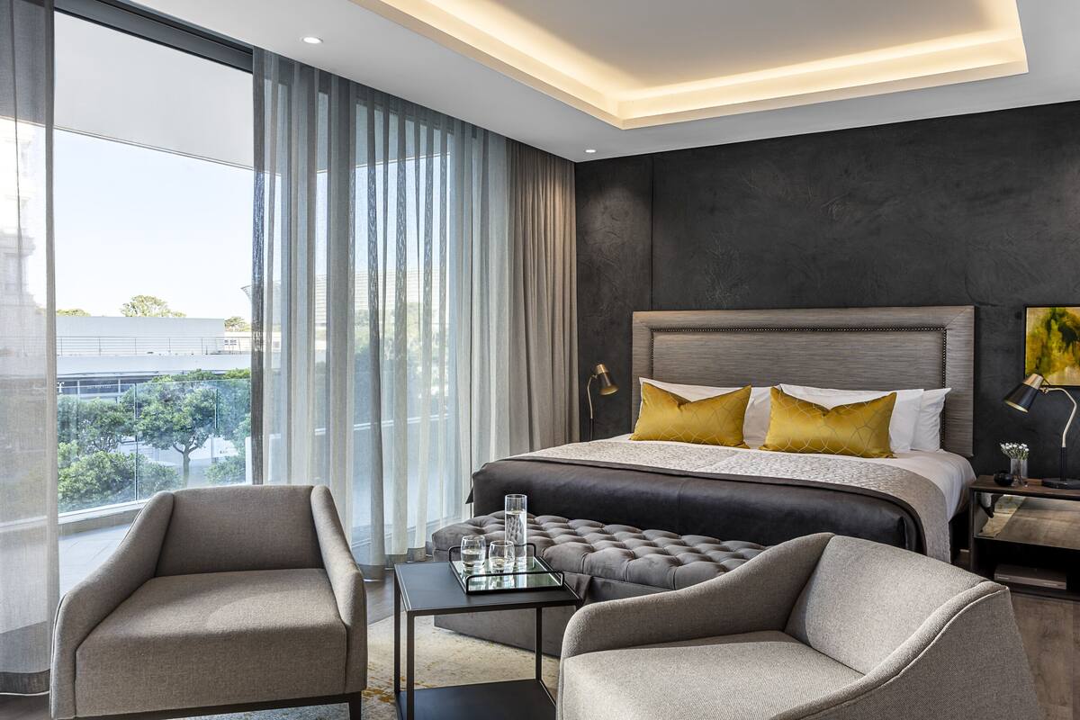 Une chambre d’hôtel moderne dotée d’un grand lit, d’un coin salon avec une table et des chaises, et offrant une vue sur la ville grâce à de grandes fenêtres.