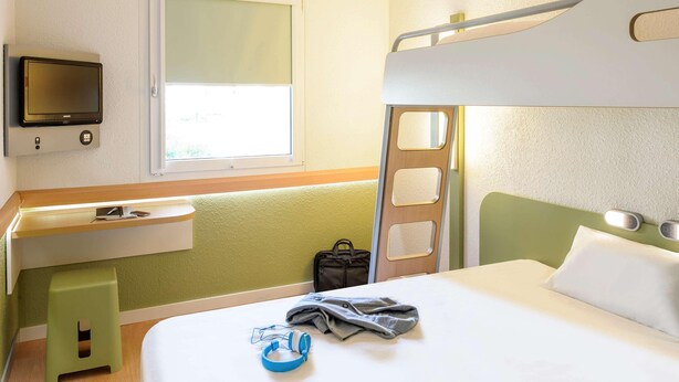 Ein Hotelzimmer mit einem Etagenbett, einem Nachttisch mit Lampe, einem an der Wand montierten Fernseher und einem Fenster, das für natürliches Licht sorgt.