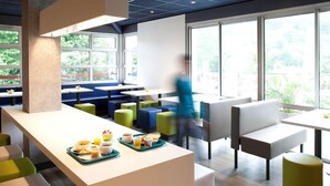 A modern dining area with high tables and booths, a central serving counter with breakfast items, and large windows offering a view of greenery.
