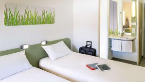 A hotel room with two beds, a bedside table with a tablet, a suitcase, and a bathroom with a sink and mirror. - Ibis Budget Biarritz Anglet (Anglet)