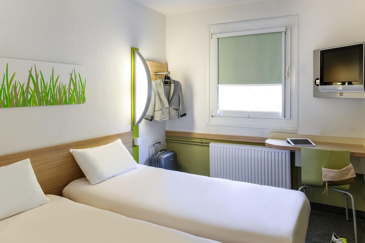 A hotel room with two beds, a desk with a computer, a window, and a radiator.