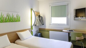 A hotel room with two beds, a desk with a computer, a window, and a radiator.