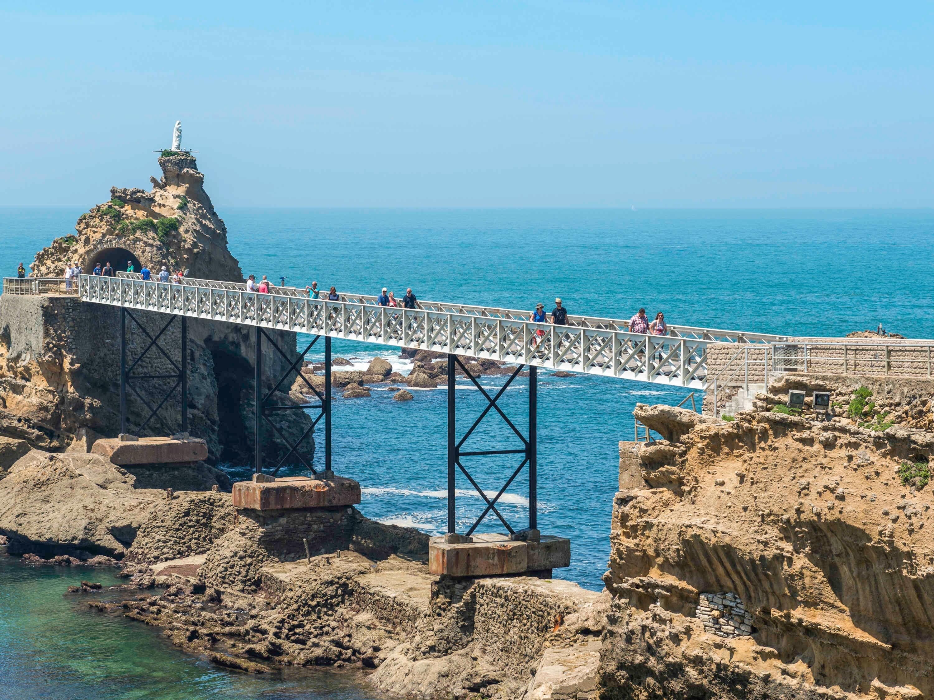 A bridge over rocky terrain with a statue on top, leading to a coastal area.