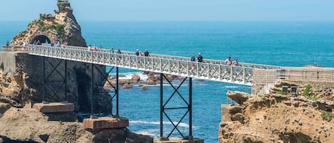 A bridge over rocky terrain with a statue on top, leading to a coastal area.