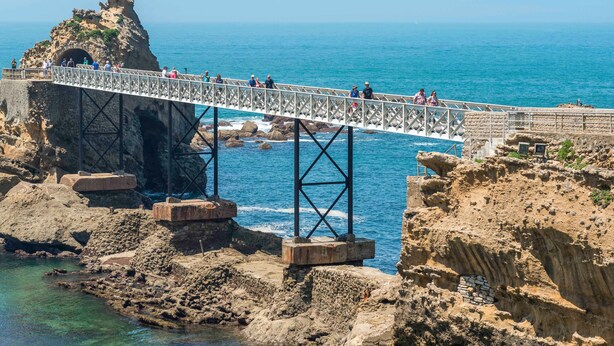 A bridge over rocky terrain with a statue on top, leading to a coastal area.