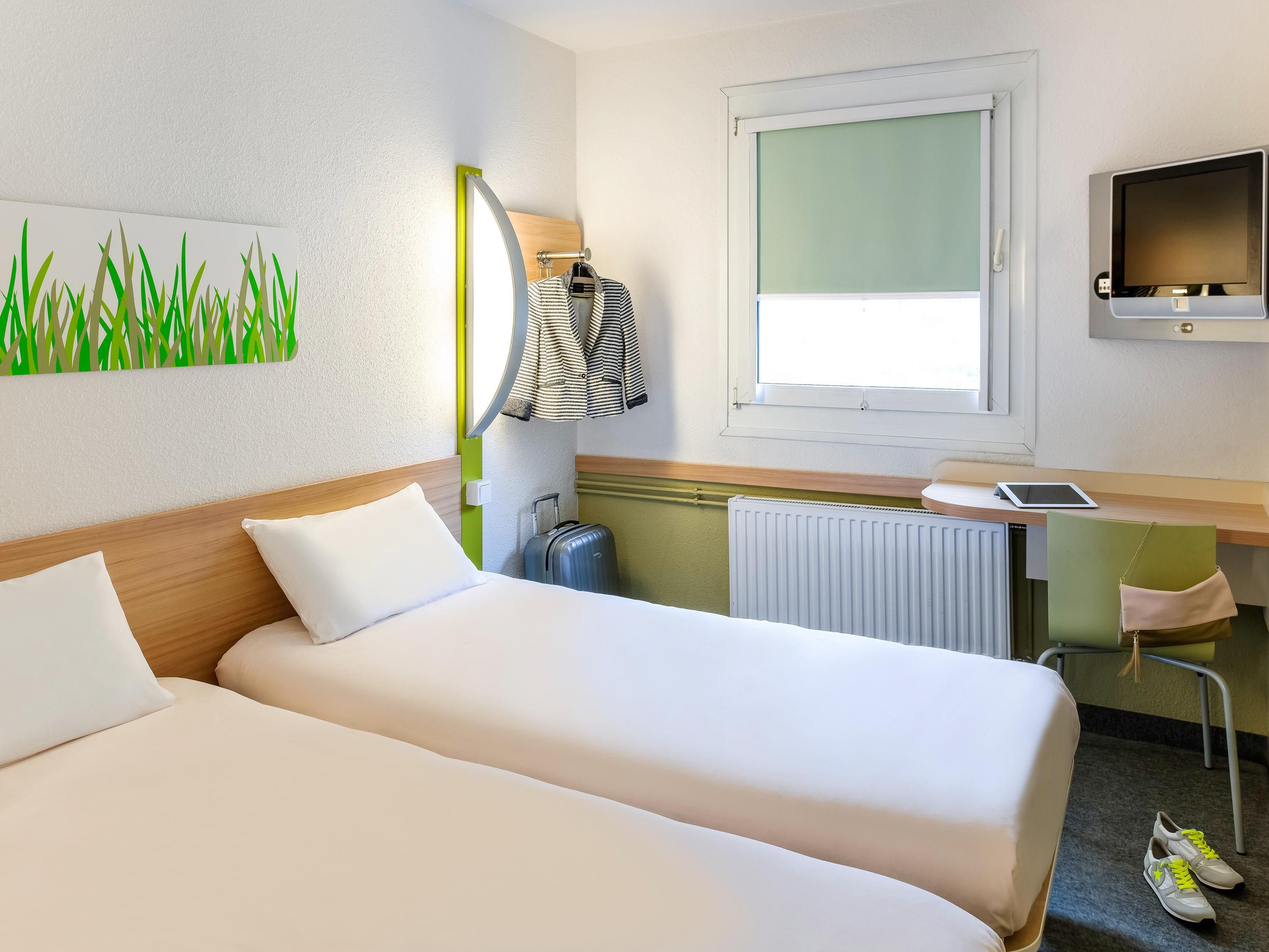 Habitación de hotel con dos camas, un escritorio con computadora, televisor, ventana y radiador.