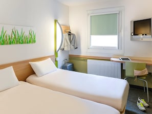 A hotel room with two beds, a desk with a computer, a TV, a window, and a radiator.