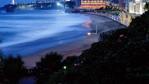 A coastal cityscape at dusk with a prominent lighthouse, a curved promenade, and a large building with illuminated windows. - Ibis Budget Biarritz Anglet (Anglet)