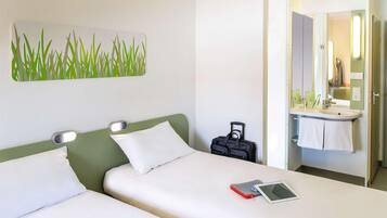 Habitación de hotel con dos camas, una mesita de noche con una tableta, una maleta, y un baño con lavamanos y espejo.