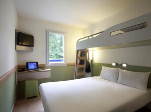 Une chambre d’hôtel compacte comprenant un lit superposé, un bureau avec un ordinateur portable, un téléviseur fixé au mur et une fenêtre donnant sur un espace verdoyant.