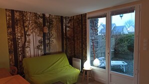 A room with a green sofa, a small stool with a lamp, and a sliding glass door leading to an outdoor area with a table and chairs. - Cottage "La Campagne" between sea and countryside (Heugueville-sur-Sienne)