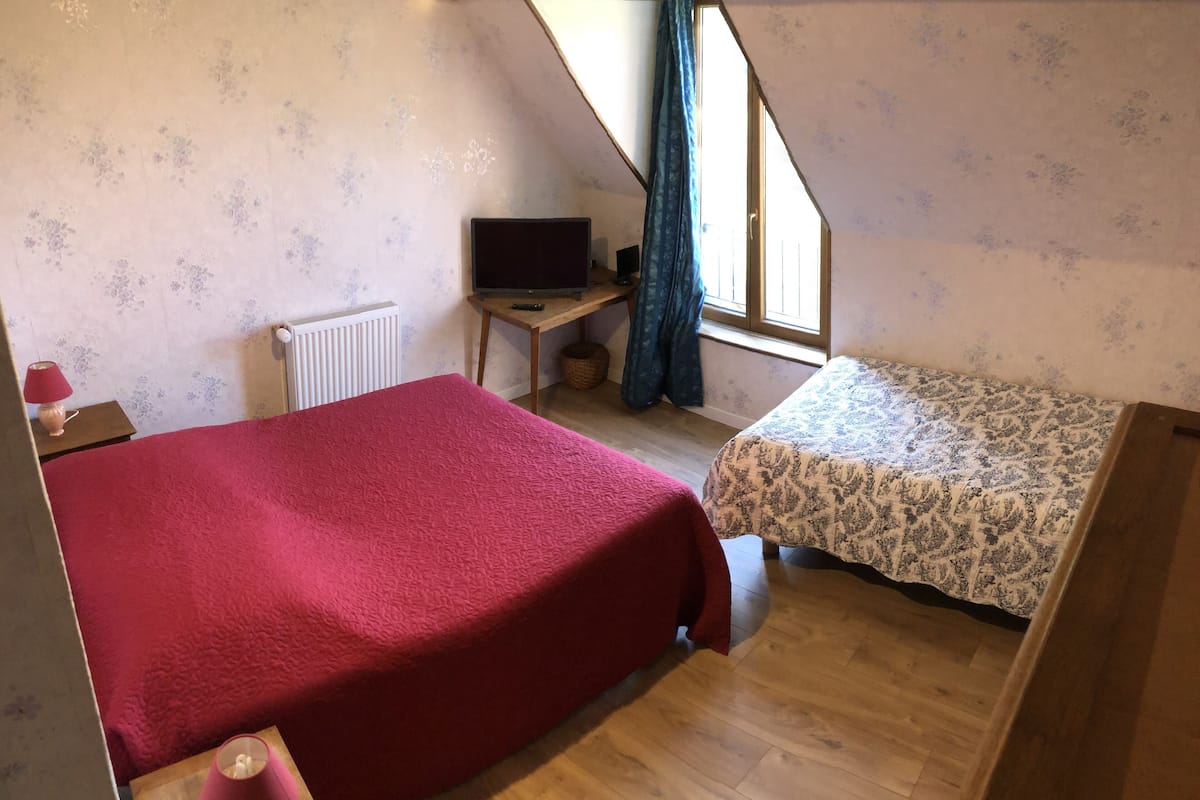 A bedroom with a large bed, a smaller bed, a desk with a television, and a window with curtains.
