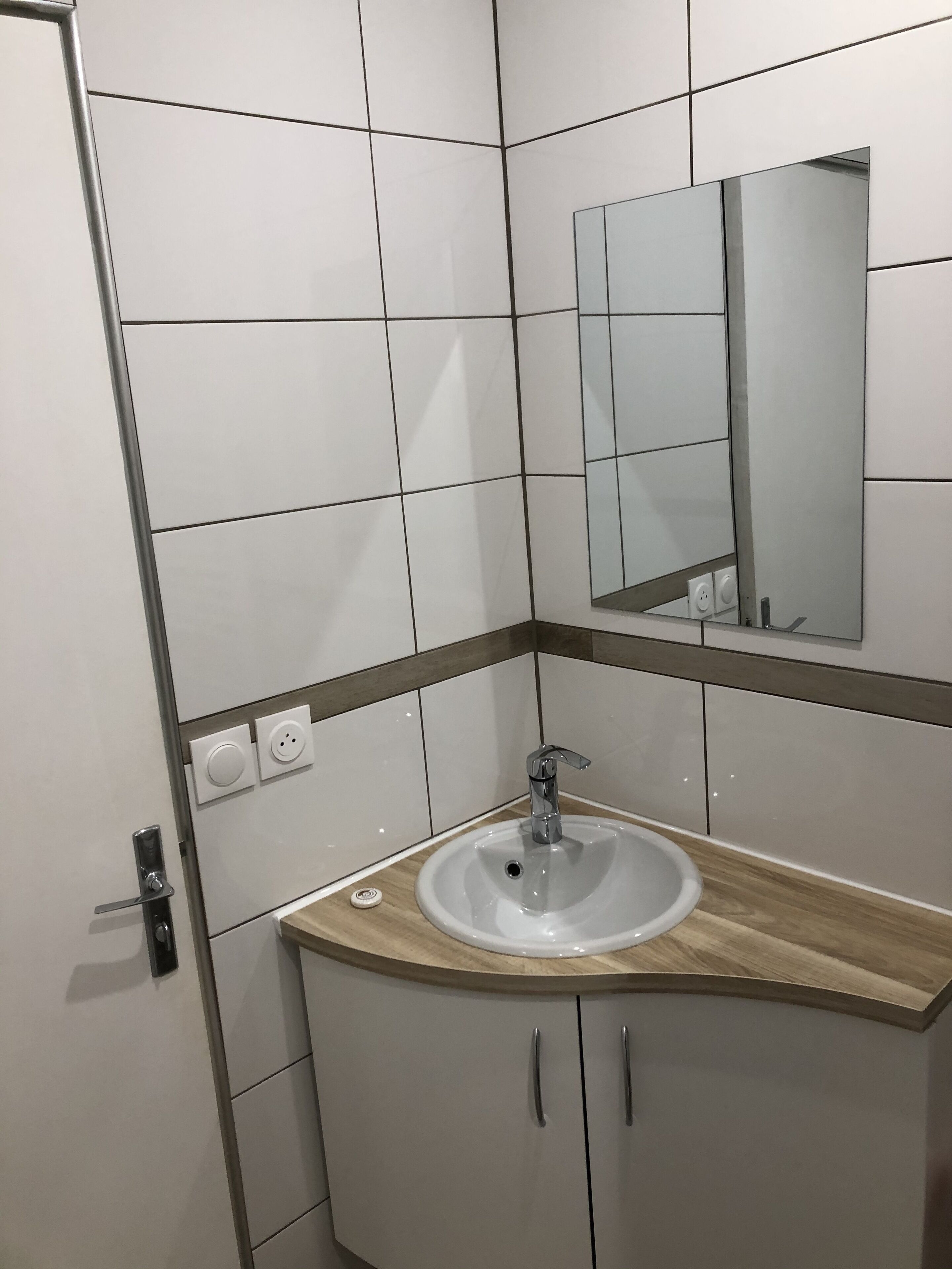 Ein Badezimmer mit weiß gefliesten Wänden, einer Holz Arbeitsplatte, einem Waschbecken mit Chromarmatur, einem Spiegel über dem Waschbecken und einer Tür mit Griff.