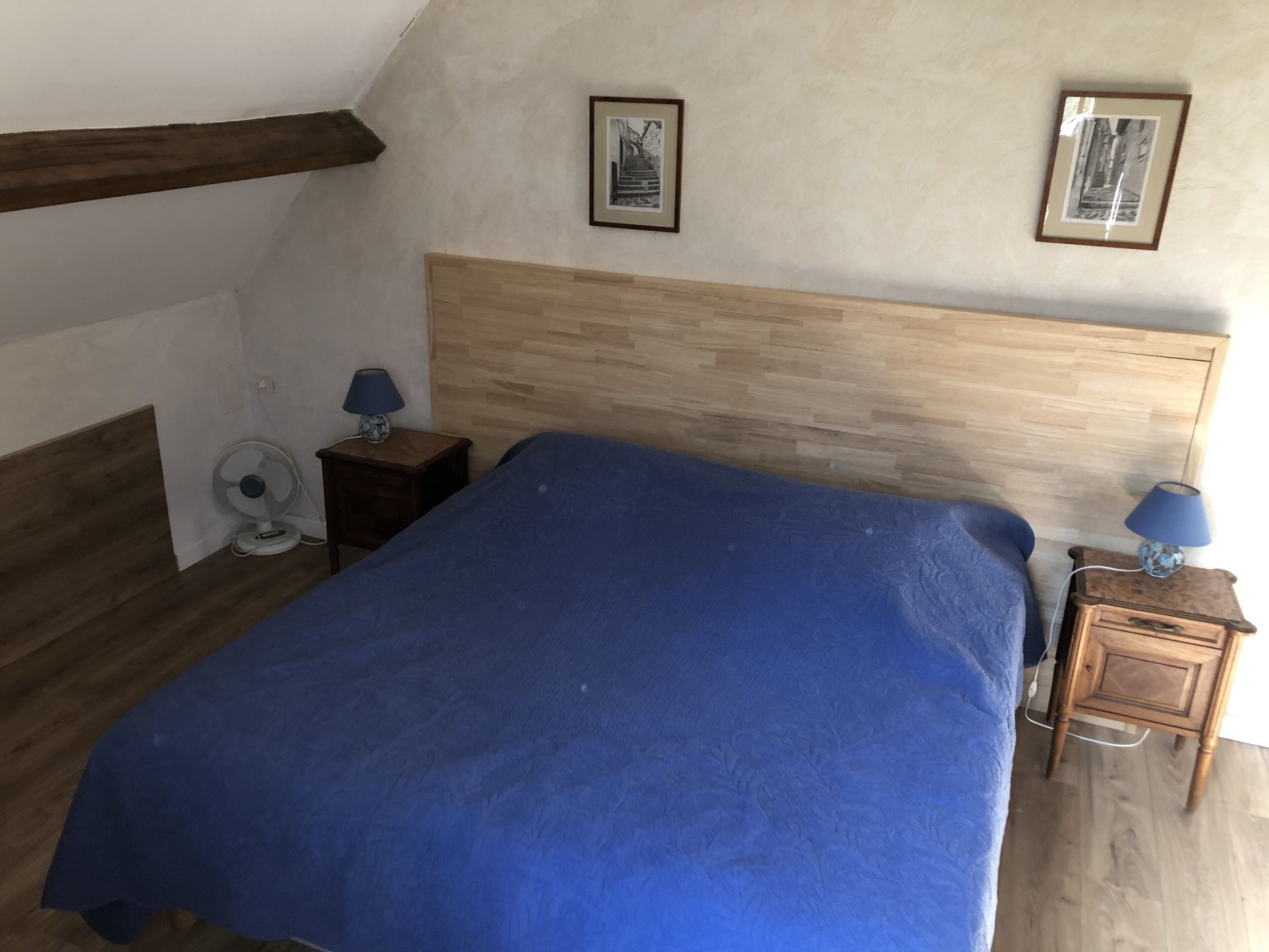 A bedroom with a wooden headboard, a bed with a blue blanket, two bedside tables with lamps, and two framed pictures on the wall.