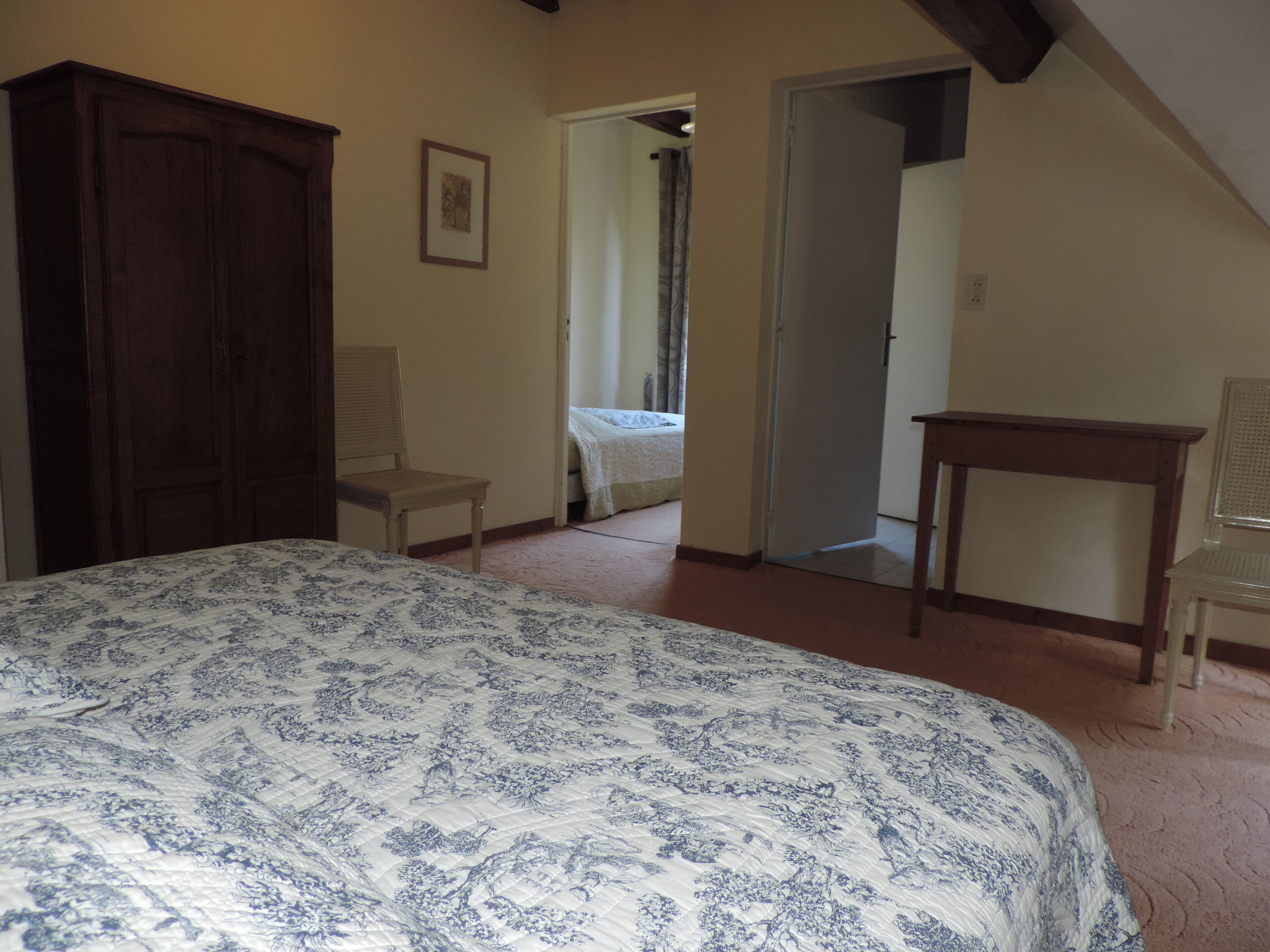 Een slaapkamer met een groot bed, een houten garderobe, een stoel en een kleine tafel. Er is een deur die naar een andere kamer leidt.