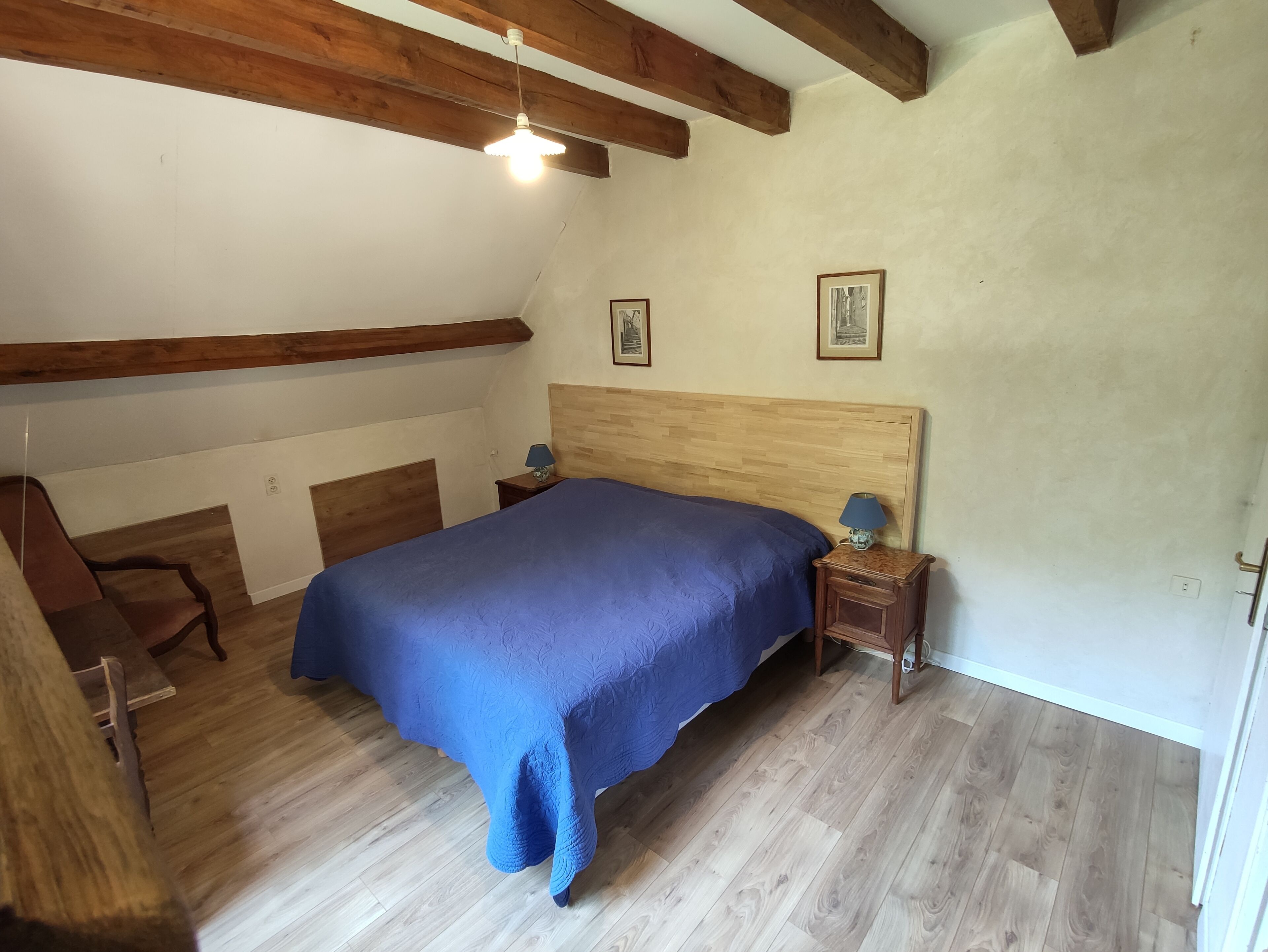 Un dormitorio con techo de madera, una cama con colcha azul, dos mesitas de noche con lámparas y dos cuadros enmarcados en la pared.