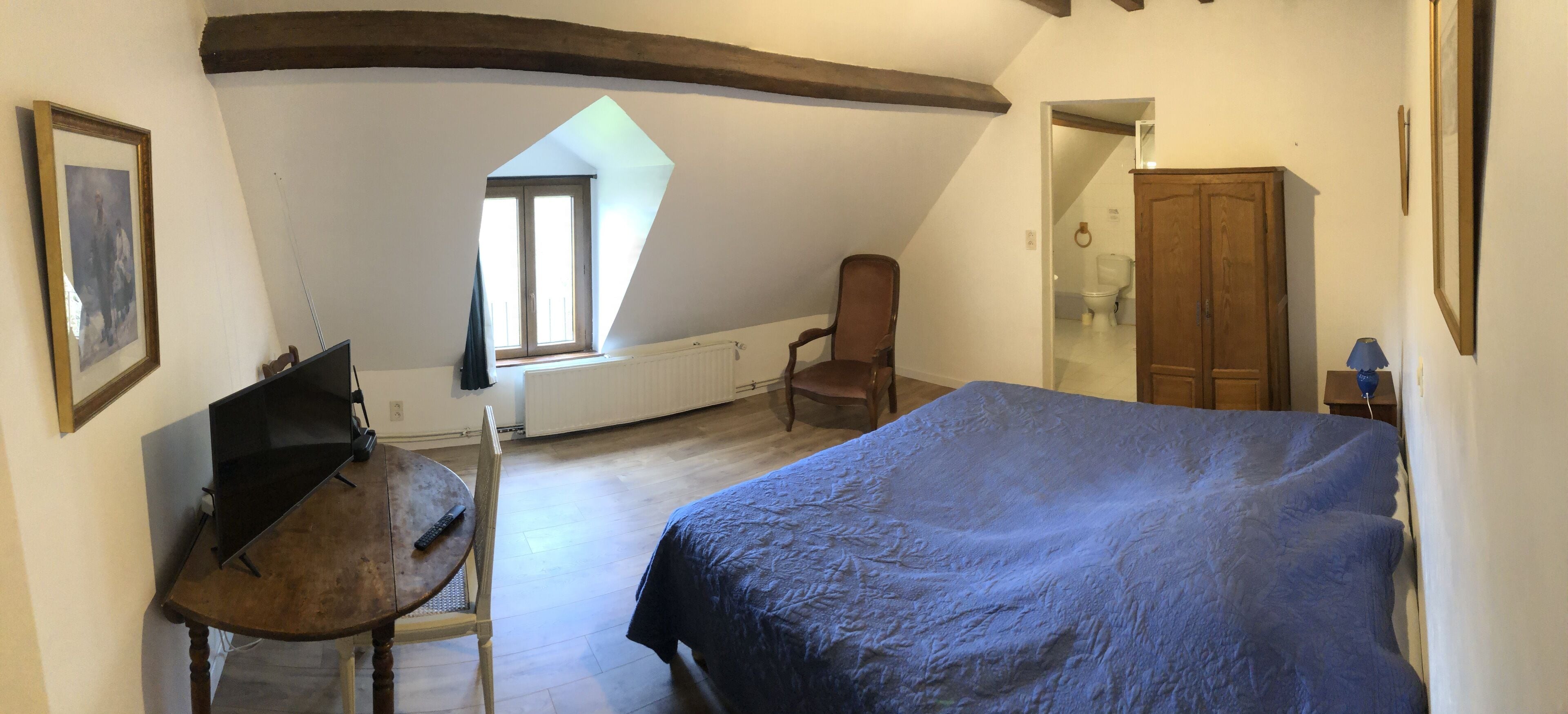 Un dormitorio con techo inclinado, cama con cubierta azul, una silla de madera, una mesa con un control remoto y una ventana con vistas al exterior.