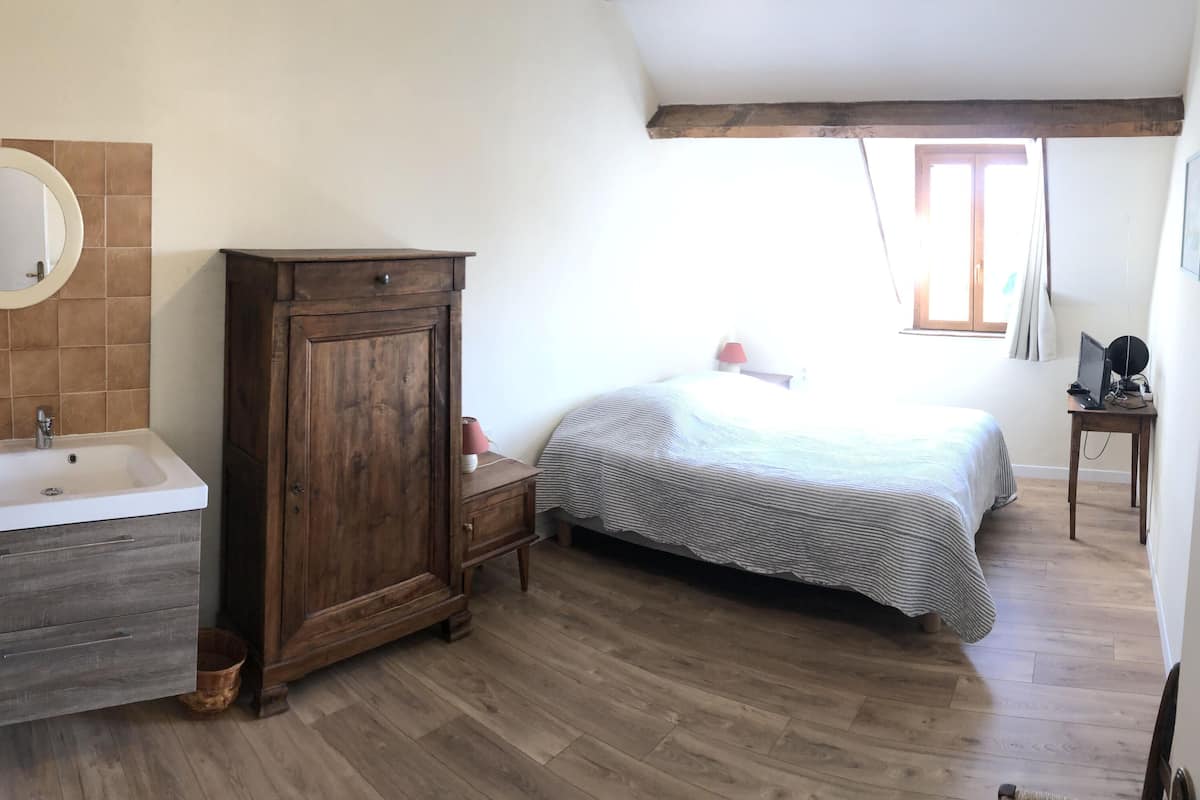 A bedroom with a wooden floor, a bed with a striped bedspread, a wooden wardrobe, a round mirror, and a white sink.