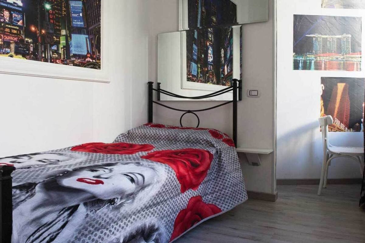 A bedroom with a bed, a window with a cityscape view, a chair, and a framed picture on the wall.