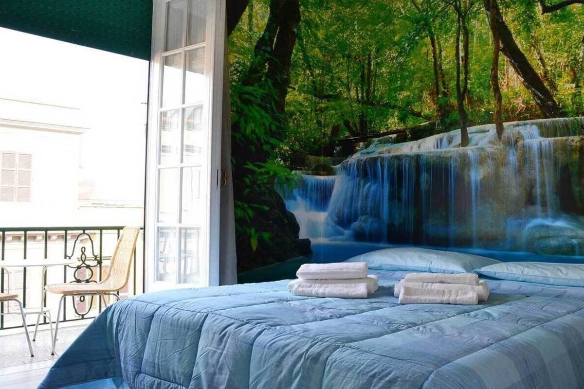 A bedroom with a large bed, a view of a waterfall mural, and a balcony with a chair.