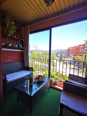 A balcony with wicker furniture, a table with a fruit bowl and wine glasses, and a view of a cityscape with buildings and greenery.