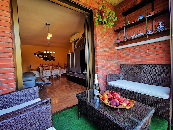 A cozy living area with wicker furniture, a glass coffee table with fruit, and a brick wall with shelves.
