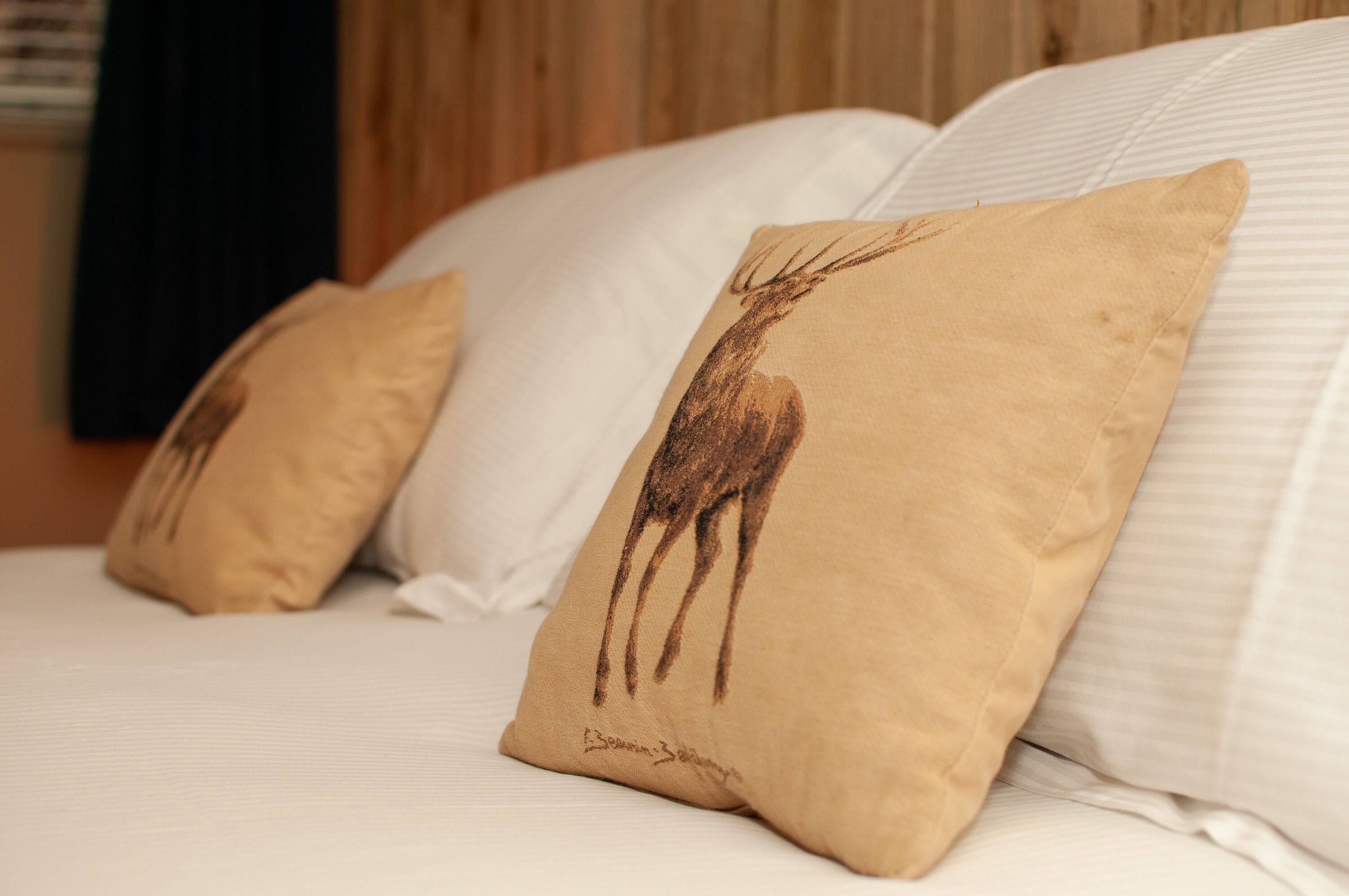 Una cama con sábanas blancas y dos almohadas color beige con ilustraciones de ciervos.
