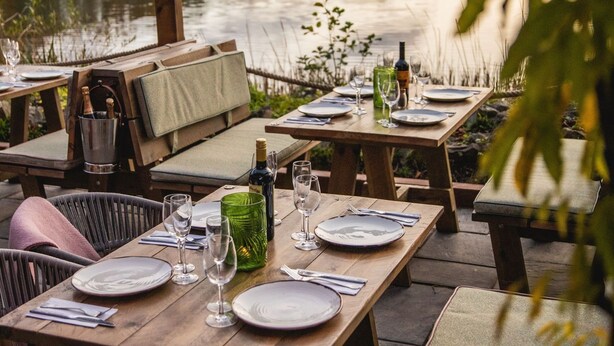 Un área de comedor al aire libre con mesas y sillas, una pérgola de madera y vistas a un lago.