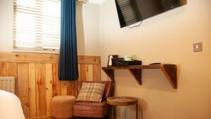 Una habitación acogedora con pared de madera, un televisor montado en la pared, una silla con cojín de cuadros, una mesita redonda y una cesta de mimbre en un estante.