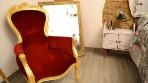 Ein Zimmer mit einem roten Samt-Sessel, einer weißen Kommode mit Spiegel und einem Bett mit gemusterten Bettwaren.