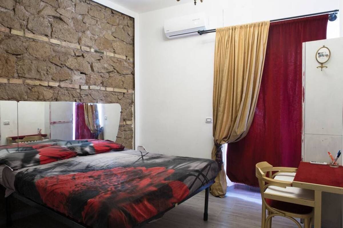 A bedroom with a stone wall, a bed with a red and black blanket, a desk with a chair, and a red curtain.