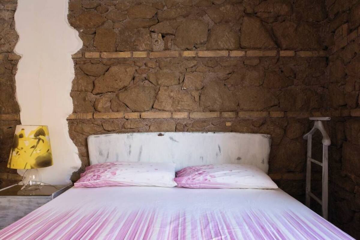A bedroom with a stone wall, a bed with white bedding, and a nightstand with a lamp.