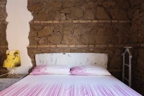 A bedroom with a stone wall, a bed with white bedding, and a nightstand with a lamp.