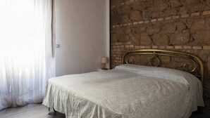 Ein Schlafzimmer mit Steinwand, einem Bett mit Metallkopfteil und einem Fenster mit durchscheinenden Vorhängen.