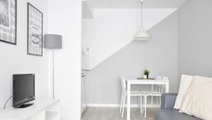 Una sala de estar moderna con un sofá gris, una mesa de comedor blanca con sillas, un televisor sobre un mueble blanco y una lámpara.