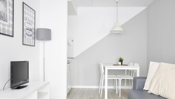 Una sala de estar moderna con un sofá gris, una mesa de comedor blanca con sillas, un televisor sobre un mueble blanco y una lámpara.