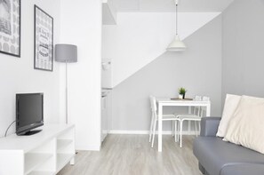 Ett modernt vardagsrum med en grå soffa, ett vitt matbord med stolar, en tv på ett vitt tv-bord och en lampa.