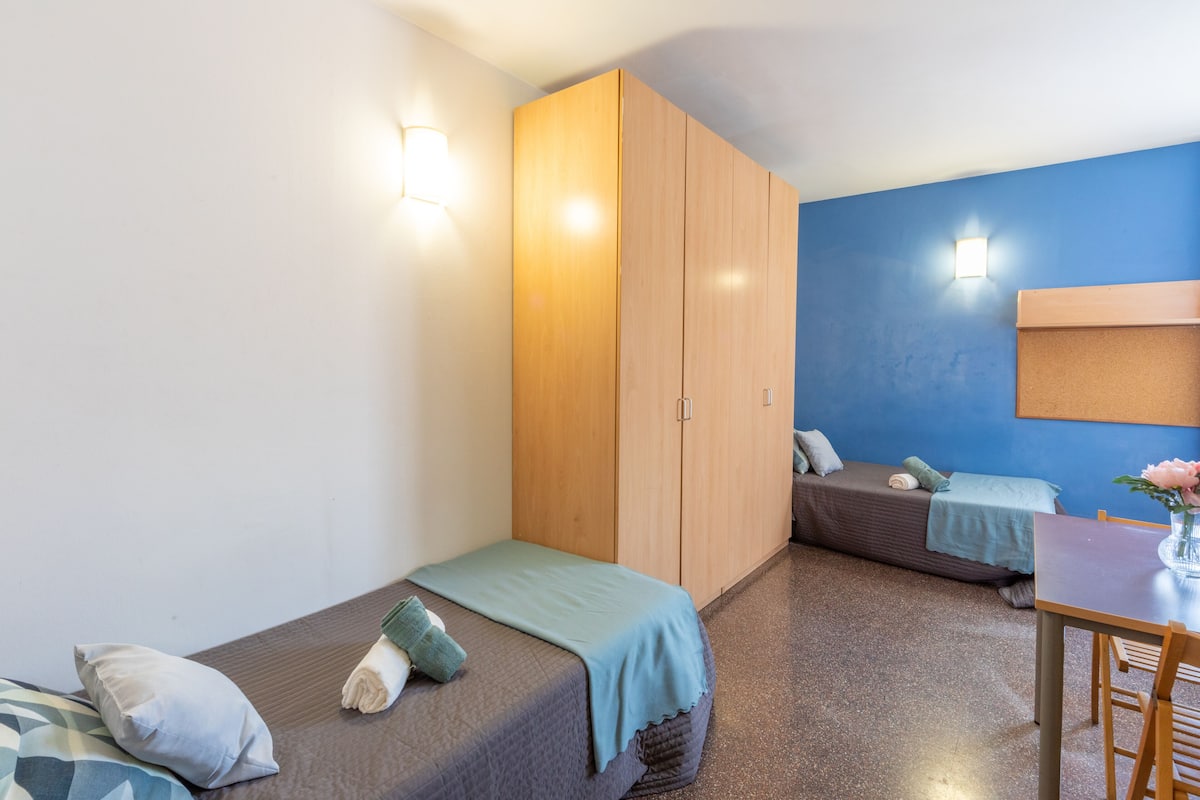 Habitación compacta con dos camas individuales, un armario de madera, una mesita pequeña y un escritorio con silla.