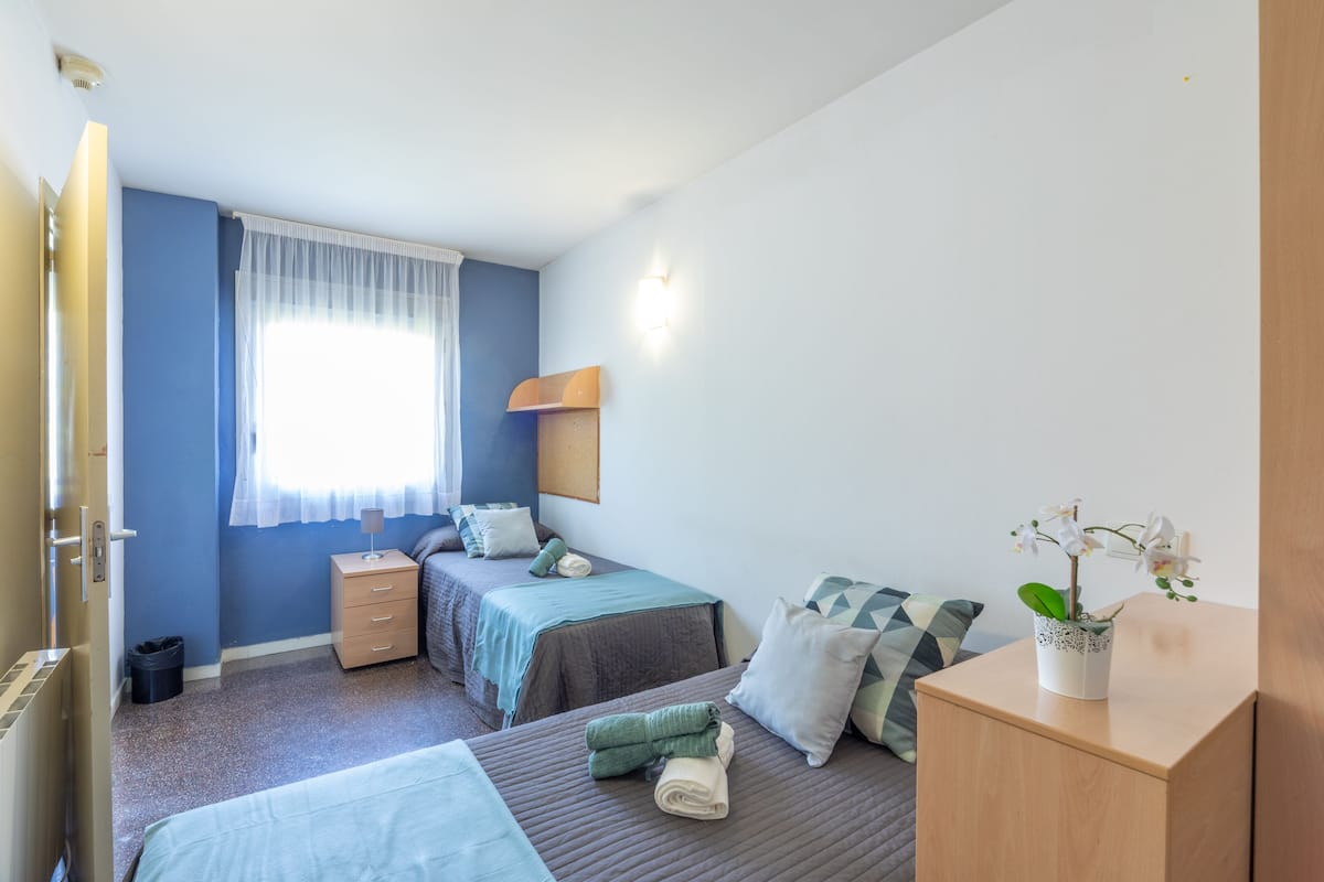 Habitación de hotel con dos camas, una mesita de noche, un armario pequeño, una ventana con cortinas traslúcidas y una lámpara fijada a la pared.